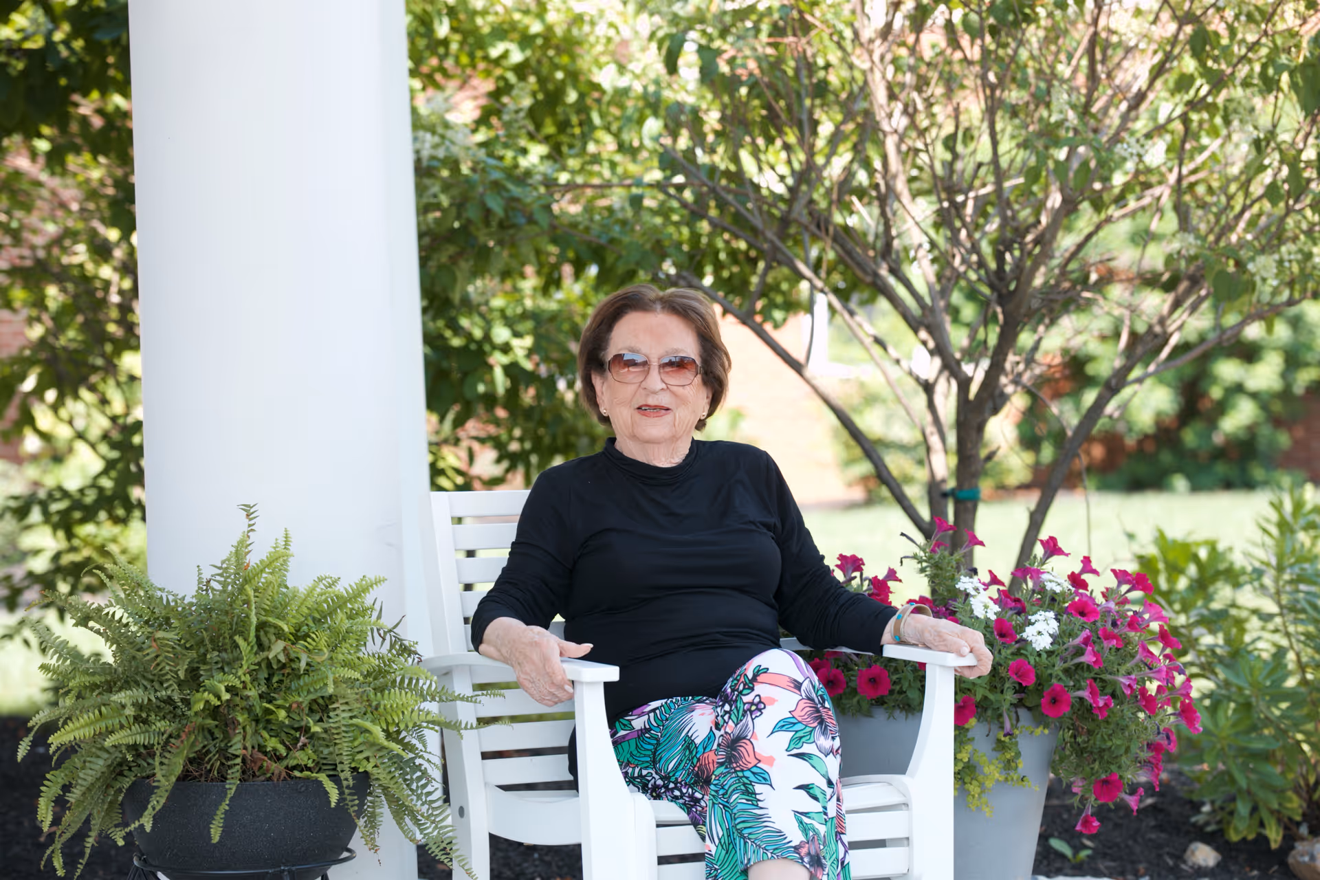 An elderly woman wearing sunglasses, a black long-sleeve top, and colorful floral pants is sitting on a white wooden chair outdoors. She is surrounded by green plants and vibrant pink and white flowers, with a tree and greenery in the background.
