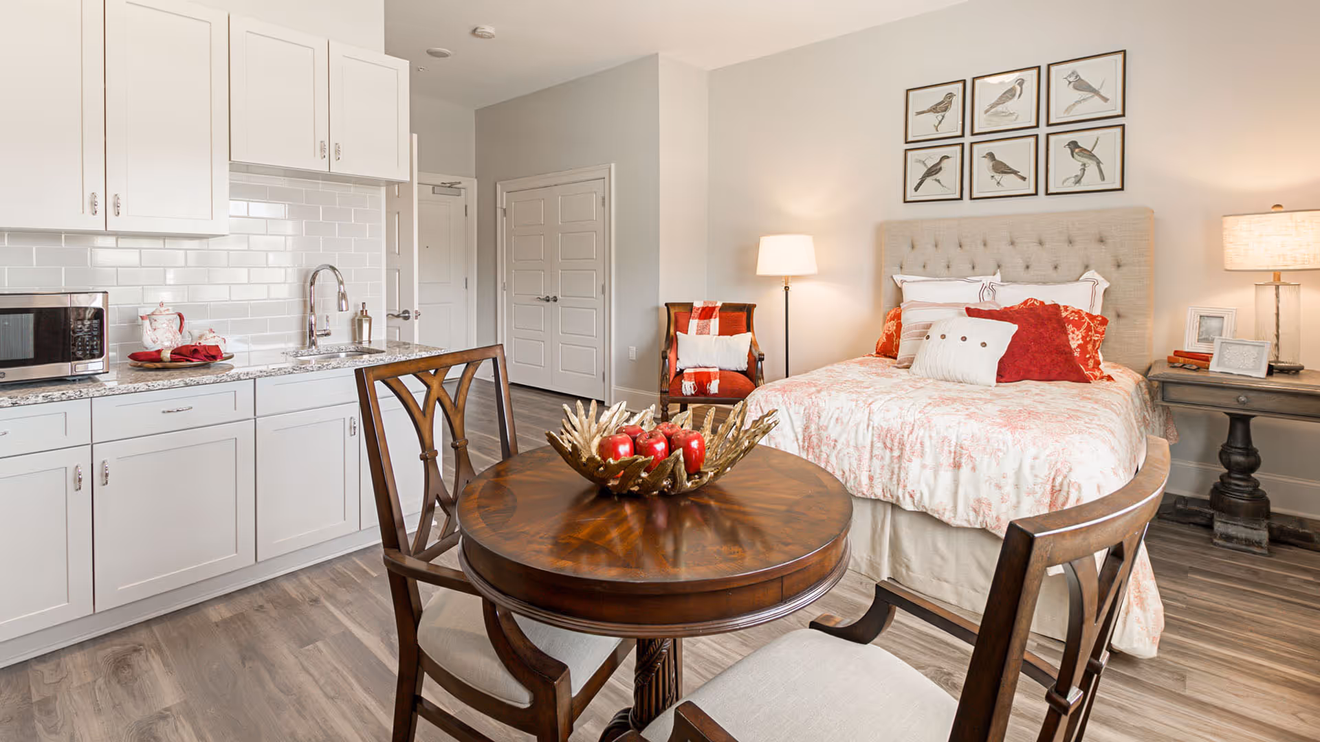 A cozy studio apartment featuring a small kitchen area with white cabinets, a microwave, and a sink. In the foreground, there is a round wooden table with two chairs and a decorative centerpiece. In the background, a bed with a tufted headboard is adorned with multiple pillows and a floral bedspread. The room also includes a side table with a lamp, a floor lamp, and framed bird artwork on the wall.