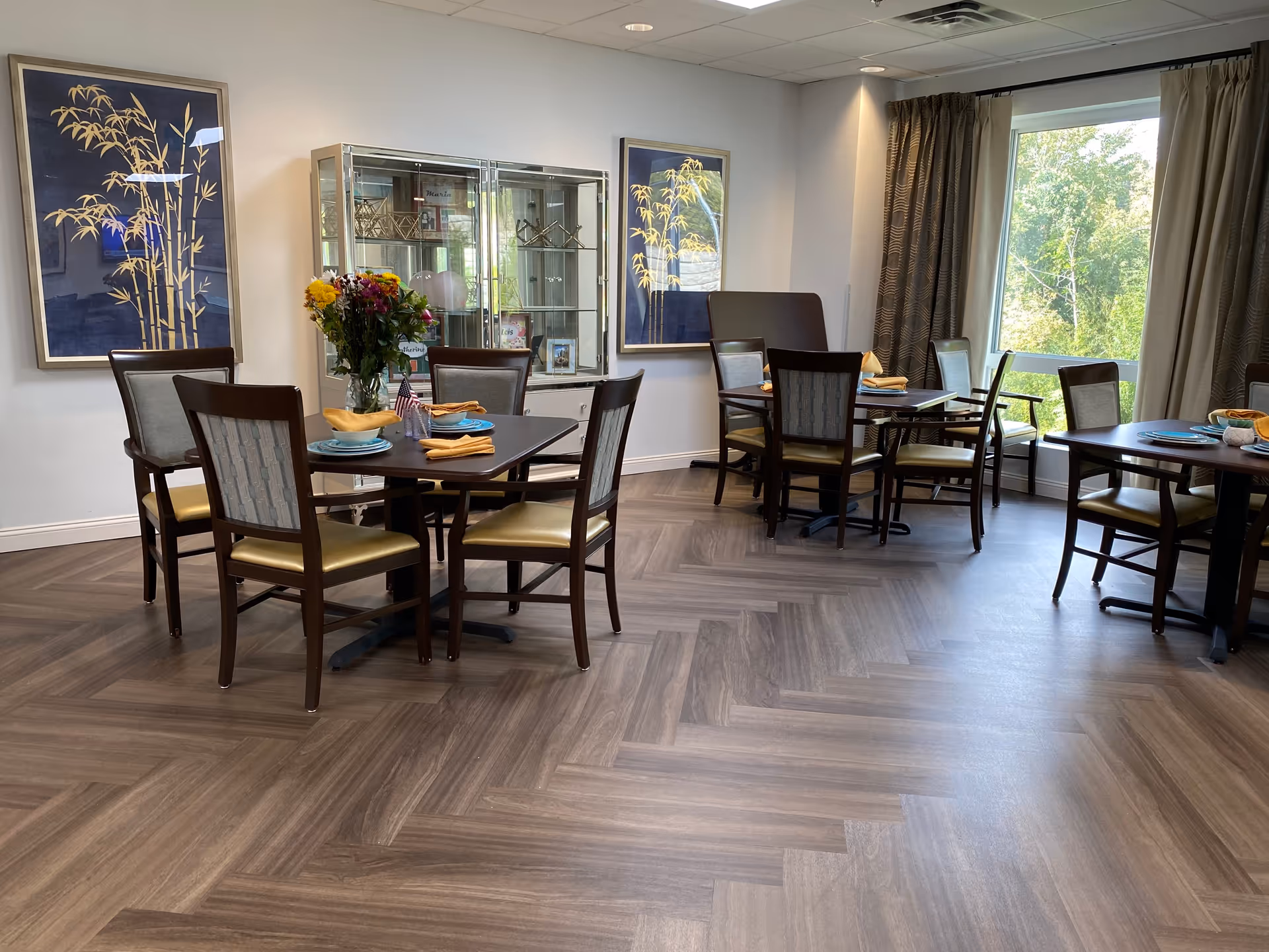 A dining room in a senior living facility with several dark wood tables and chairs arranged neatly. Each table is set with plates, napkins, and some have small American flags and a vase with flowers. The room has large windows with curtains allowing natural light to brighten the space. Two framed artworks featuring bamboo designs hang on the walls, and a glass display cabinet is visible in the background.