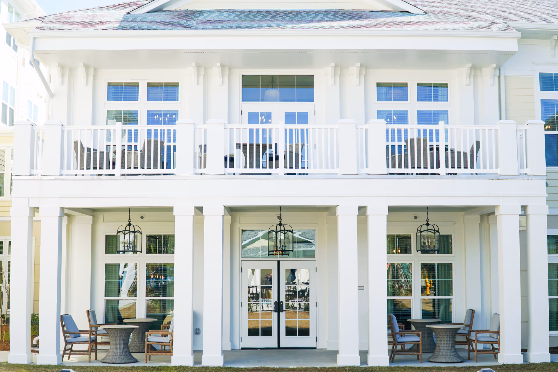 Exterior view of a white two-story building with a covered patio area on the ground floor featuring round tables and chairs, and a balcony on the upper floor with additional seating. The building has large windows and glass doors, with hanging lantern-style lights on the patio ceiling.