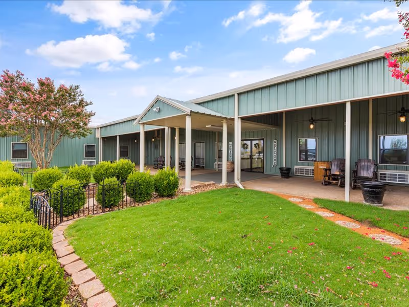 Exterior view of a single-story green building with a covered porch area supported by white columns. The porch has rocking chairs and welcome signs near the entrance. The foreground features a well-maintained green lawn, a curved stone pathway, and neatly trimmed bushes. A flowering tree is visible on the left side under a partly cloudy sky.