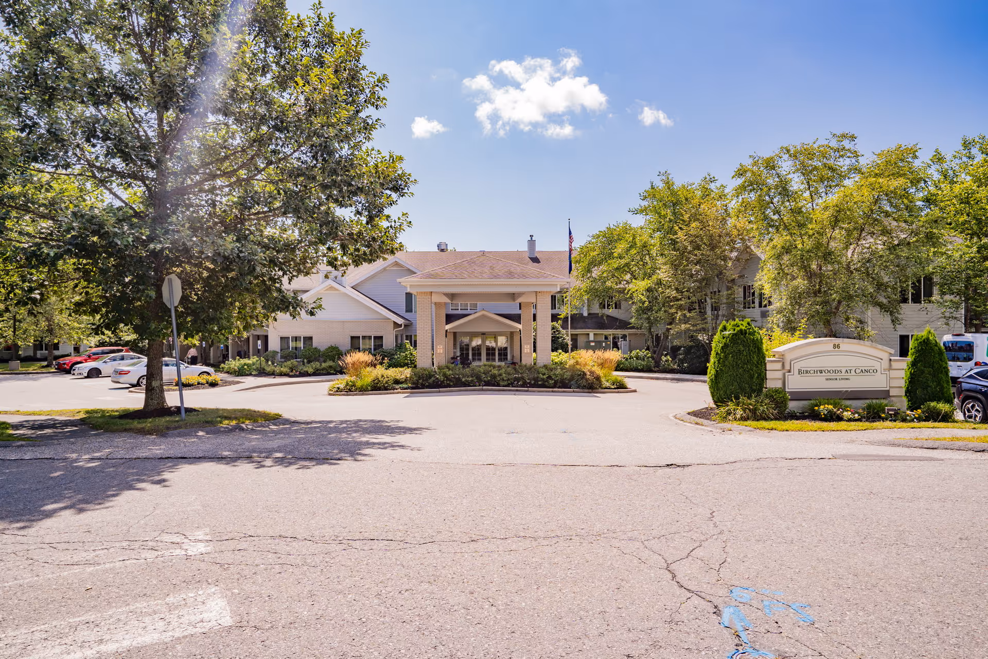Front exterior view of Birchwoods at Canco Senior Living facility with a driveway, landscaped greenery, trees, parked cars, and a clear blue sky with a few clouds.