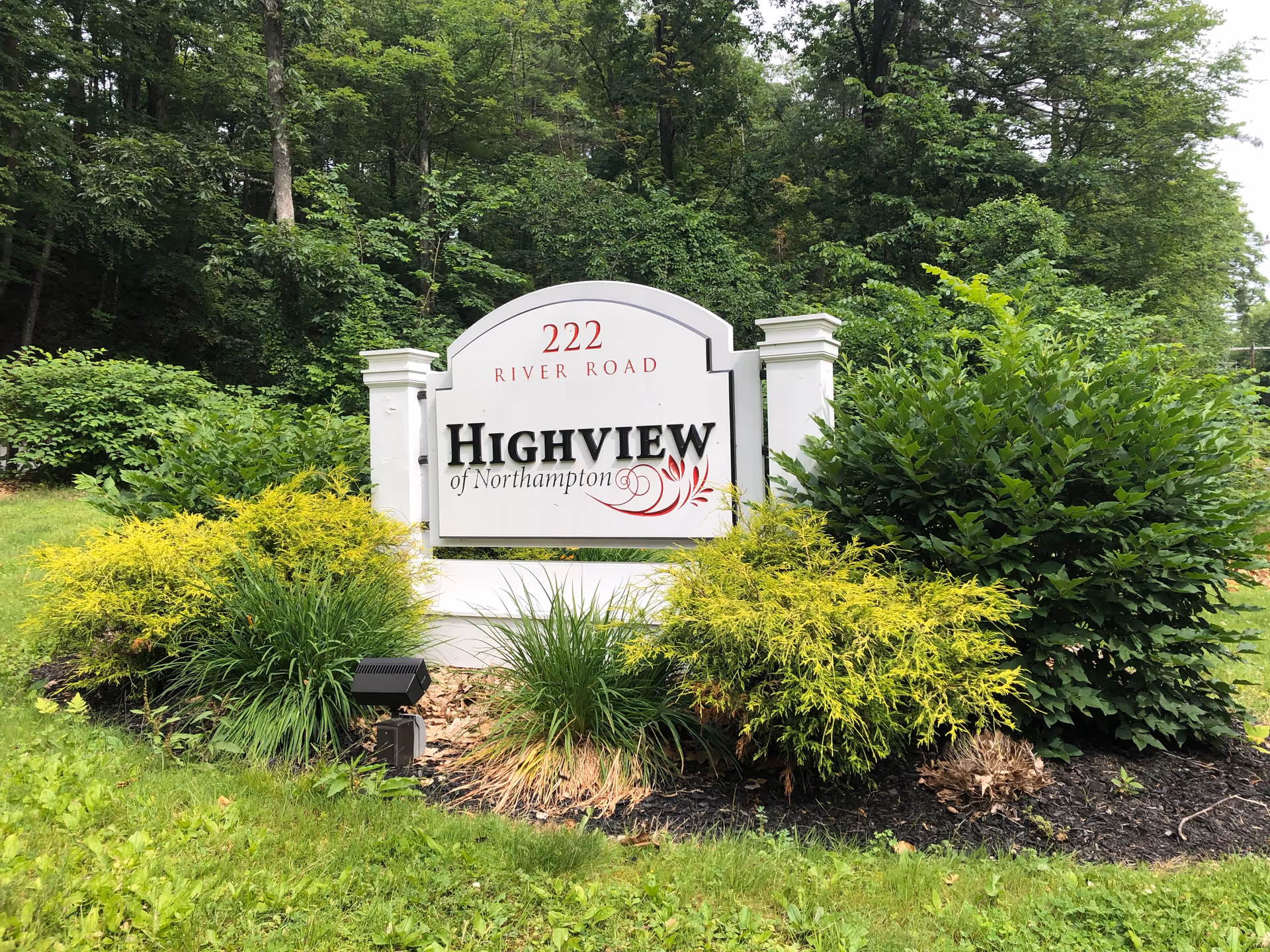 A white sign with black and red lettering that reads '222 River Road Highview of Northampton' surrounded by green bushes and trees in a landscaped area.