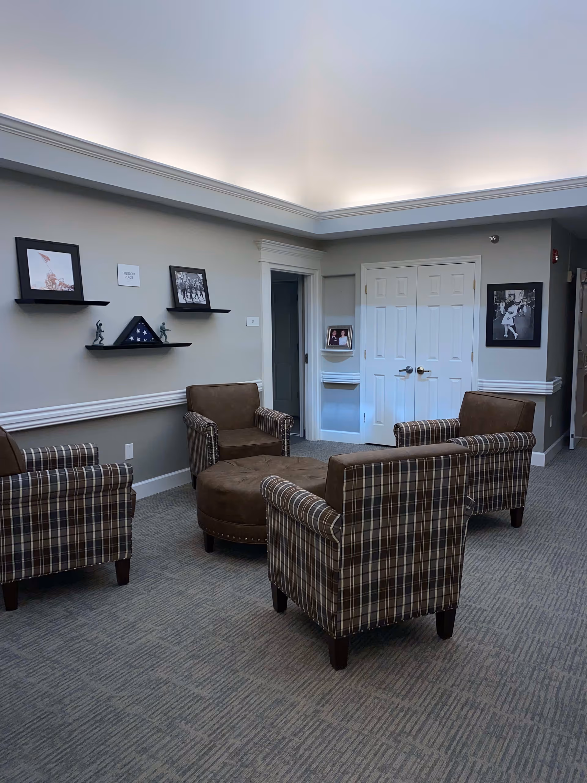 A cozy sitting area with four plaid armchairs arranged around a round brown ottoman in the center. The walls are light gray with white trim, decorated with framed pictures and shelves holding small statues and a folded American flag. There is a double white door and a small alcove with a photo on a shelf. The ceiling has recessed lighting creating a soft glow.