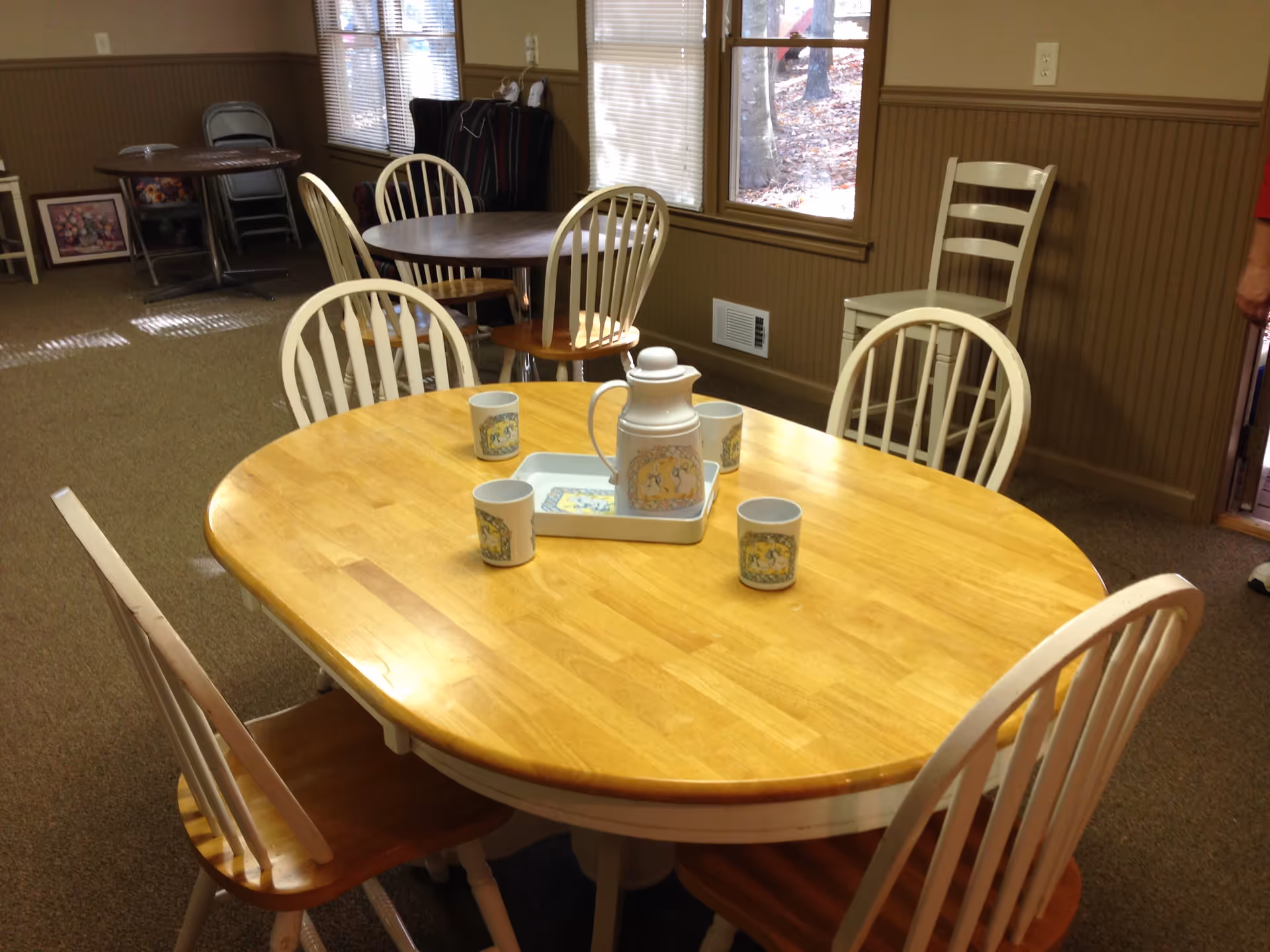 A dining area with a wooden oval table surrounded by six white chairs with wooden seats. On the table, there is a white tray holding a white pitcher and four matching mugs. In the background, there are additional tables and chairs, windows with blinds, and some folded chairs stacked against the wall.