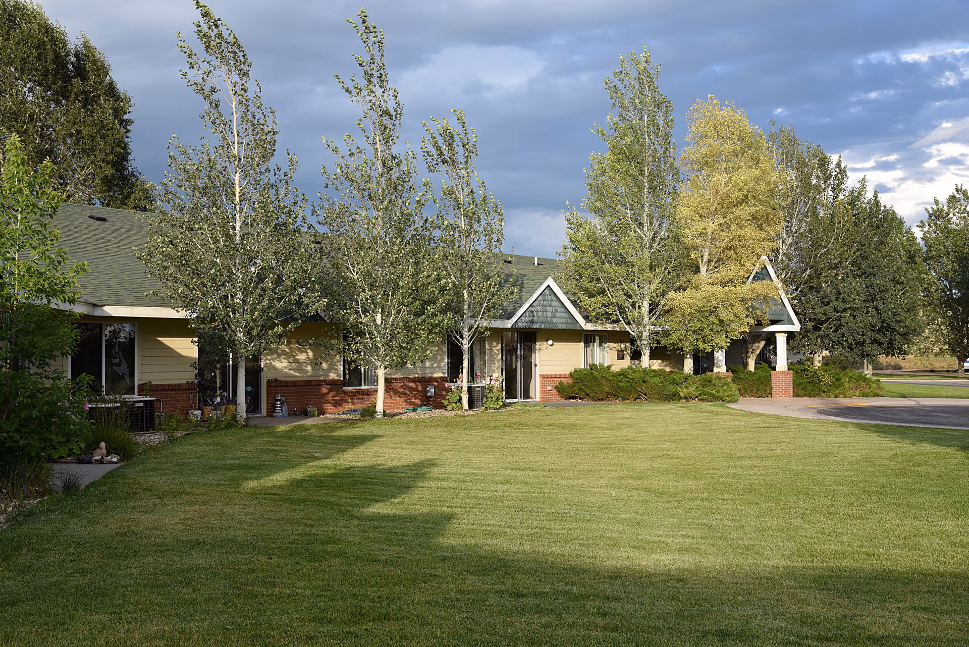 Exterior view of Regency Retirement Residence showing a single-story building with a green roof, beige and brick walls, surrounded by trees and a well-maintained lawn under a cloudy sky.