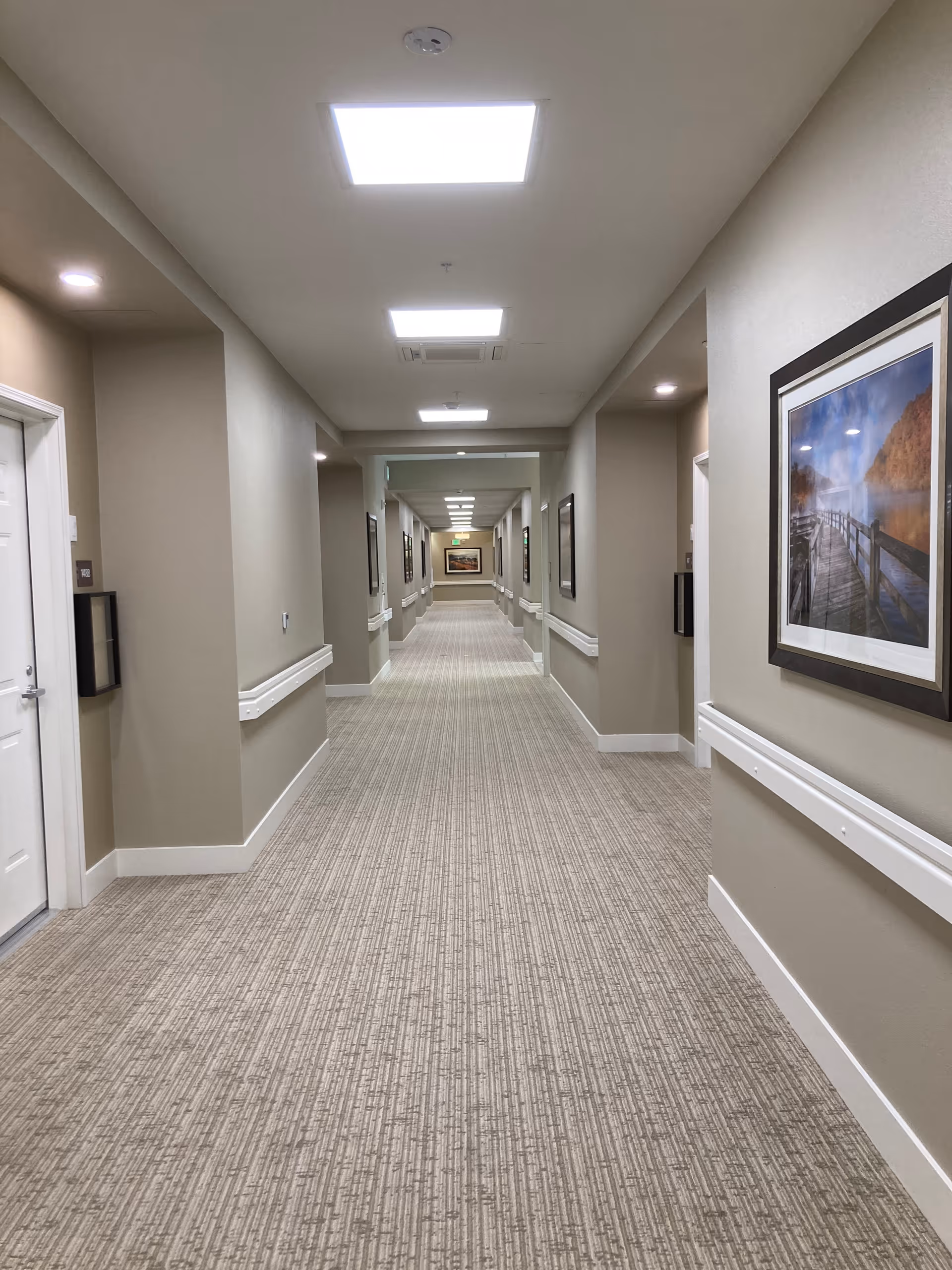 A long, well-lit hallway in an assisted living facility with beige walls, carpeted floor, white doors, handrails along the walls, and framed landscape pictures hanging on the walls.