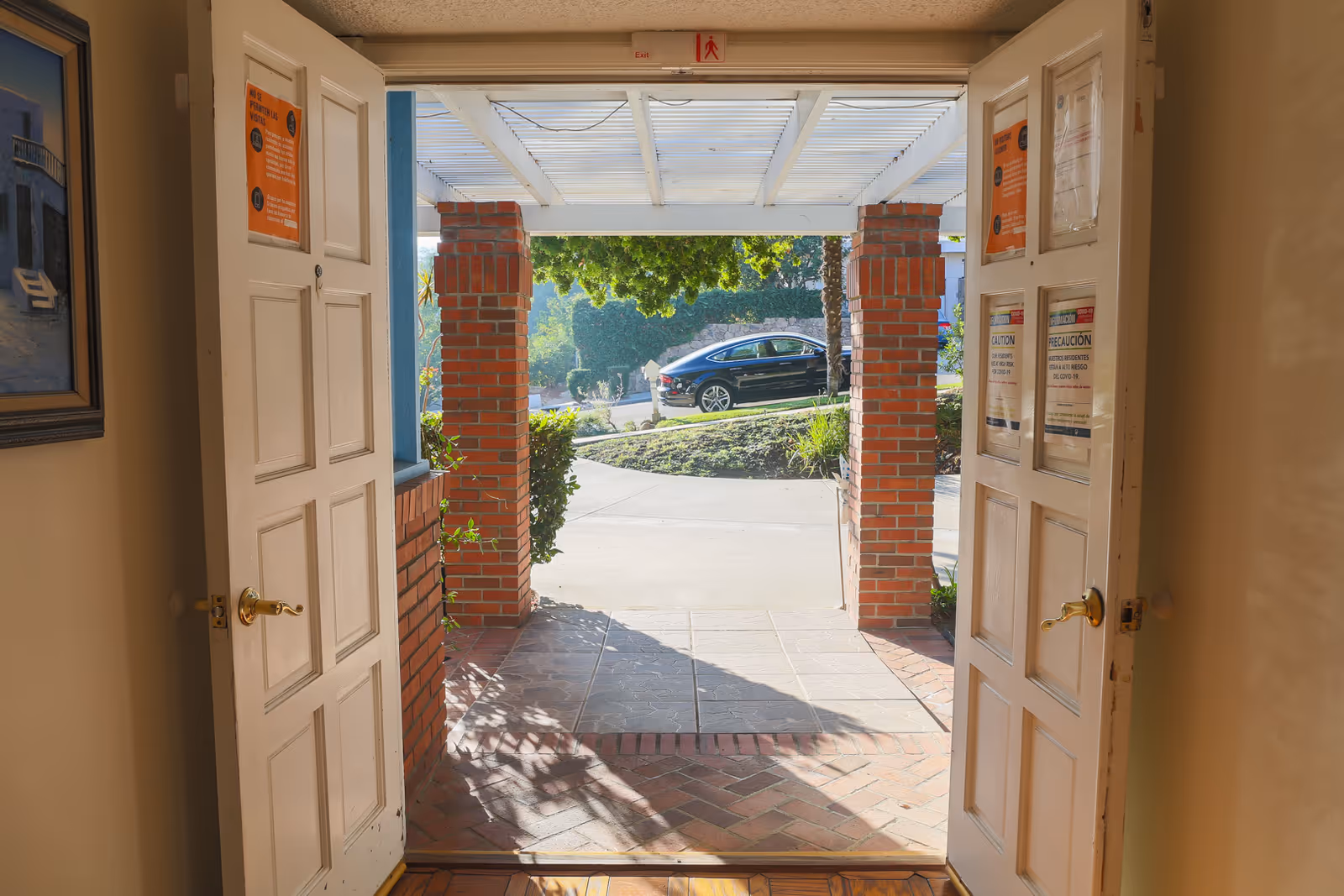 Open double doors opening onto a covered brick-pillared porch and driveway with a parked car and landscaping beyond.
