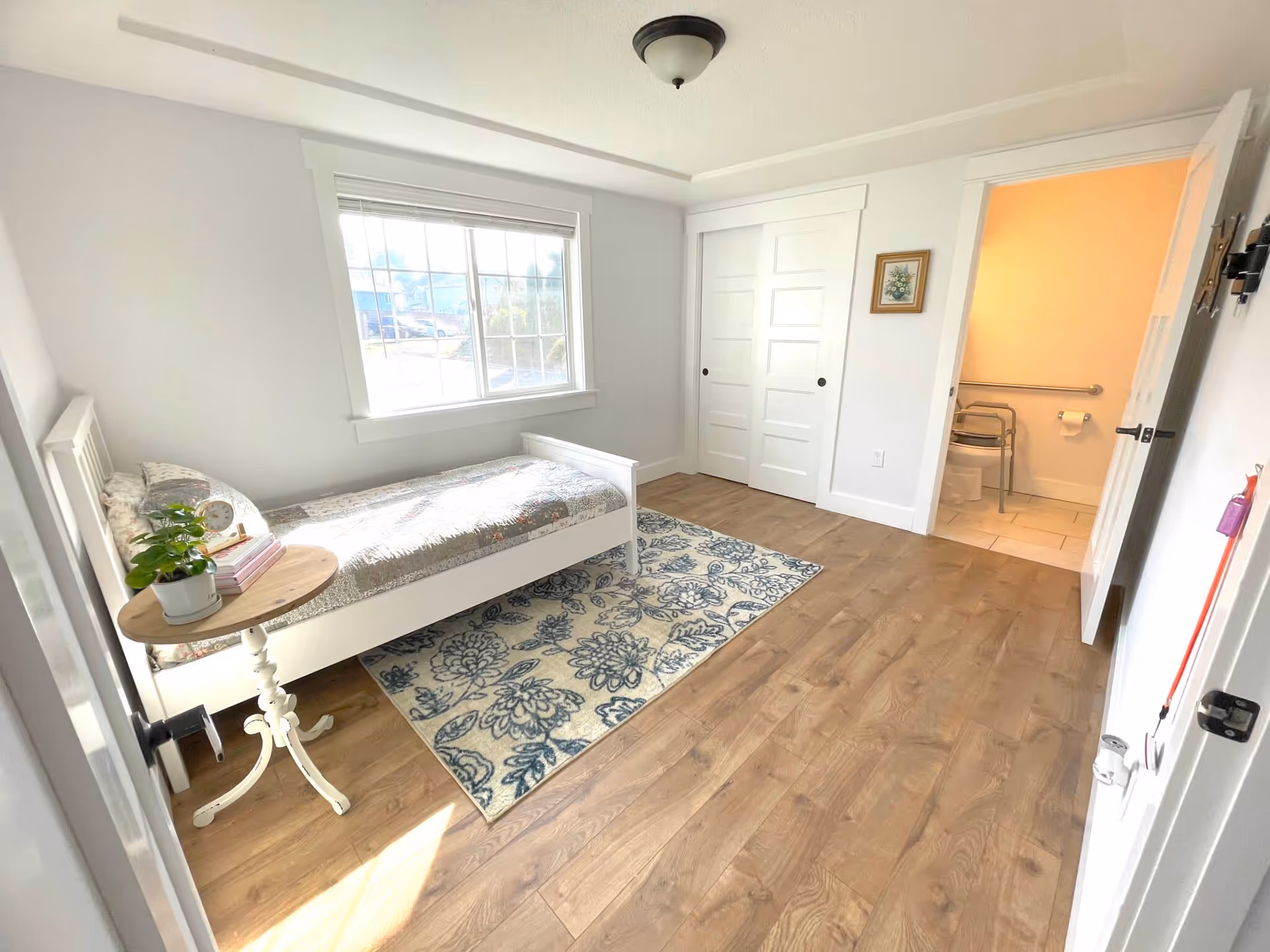 A bright bedroom with a single white bed covered with a floral quilt, a small round wooden side table with a potted plant and books, a large window letting in natural light, a blue and white floral rug on wooden flooring, white walls, and a door leading to a bathroom with a walker and grab bars visible.