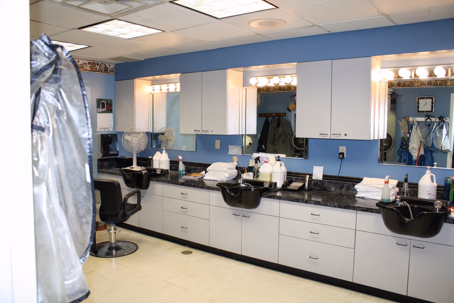 Interior view of a hair salon area in a senior living facility with three black salon sinks and chairs, a countertop with various hair care products and towels, large mirrors with lights above, and blue walls.