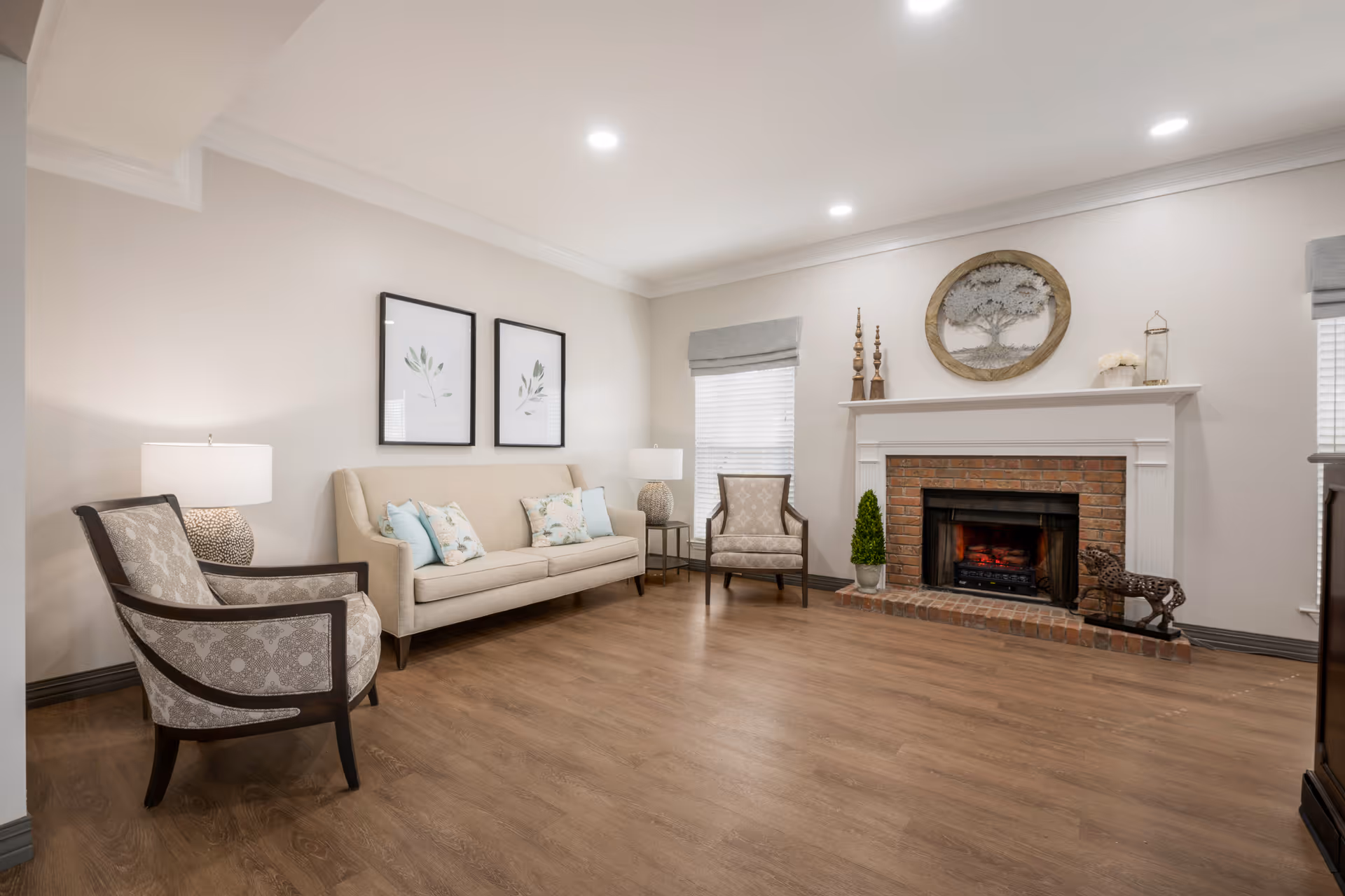 A cozy living room with a beige sofa adorned with floral and solid light blue cushions, two patterned armchairs, a brick fireplace with a decorative tree artwork above it, two windows with gray Roman shades, and wooden flooring. The room is softly lit with ceiling lights and table lamps.