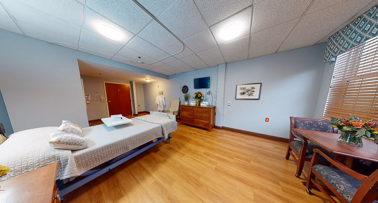 A spacious patient bedroom with a hospital bed, dresser and TV, and a seating area with table and chairs near a window.
