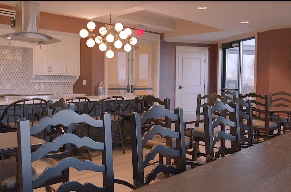 Dining room with rows of wooden chairs and tables facing a kitchen island and a modern chandelier.