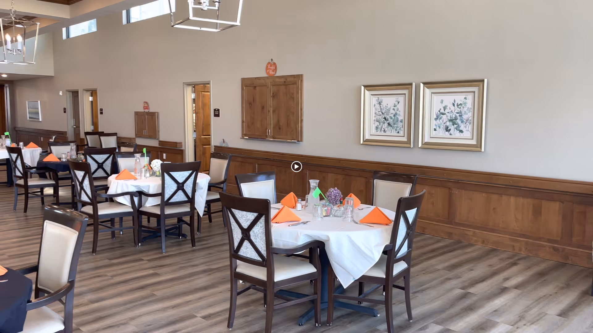 Bright dining room with round tables set with white tablecloths and orange napkins, surrounded by wooden chairs and framed artwork on the wall.