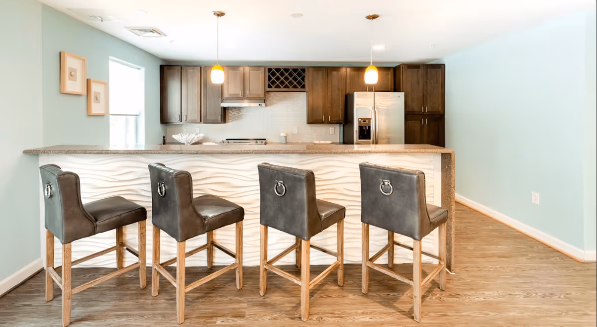 Modern kitchen with a light blue wall, wooden cabinets, stainless steel refrigerator, and a kitchen island with a textured white front panel. Four dark leather bar stools with wooden legs are lined up at the island. Two pendant lights hang above the island, and there are two framed pictures on the wall near a window.