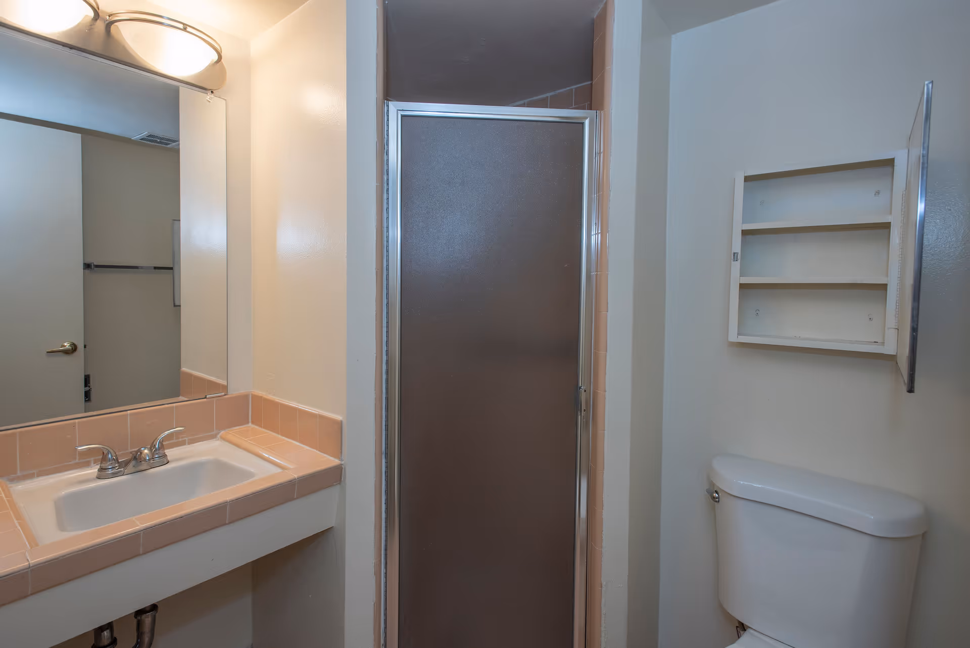 A small bathroom featuring a white toilet, an open medicine cabinet with shelves above the toilet, a sink with a pink tiled countertop, and a shower with a frosted glass door.