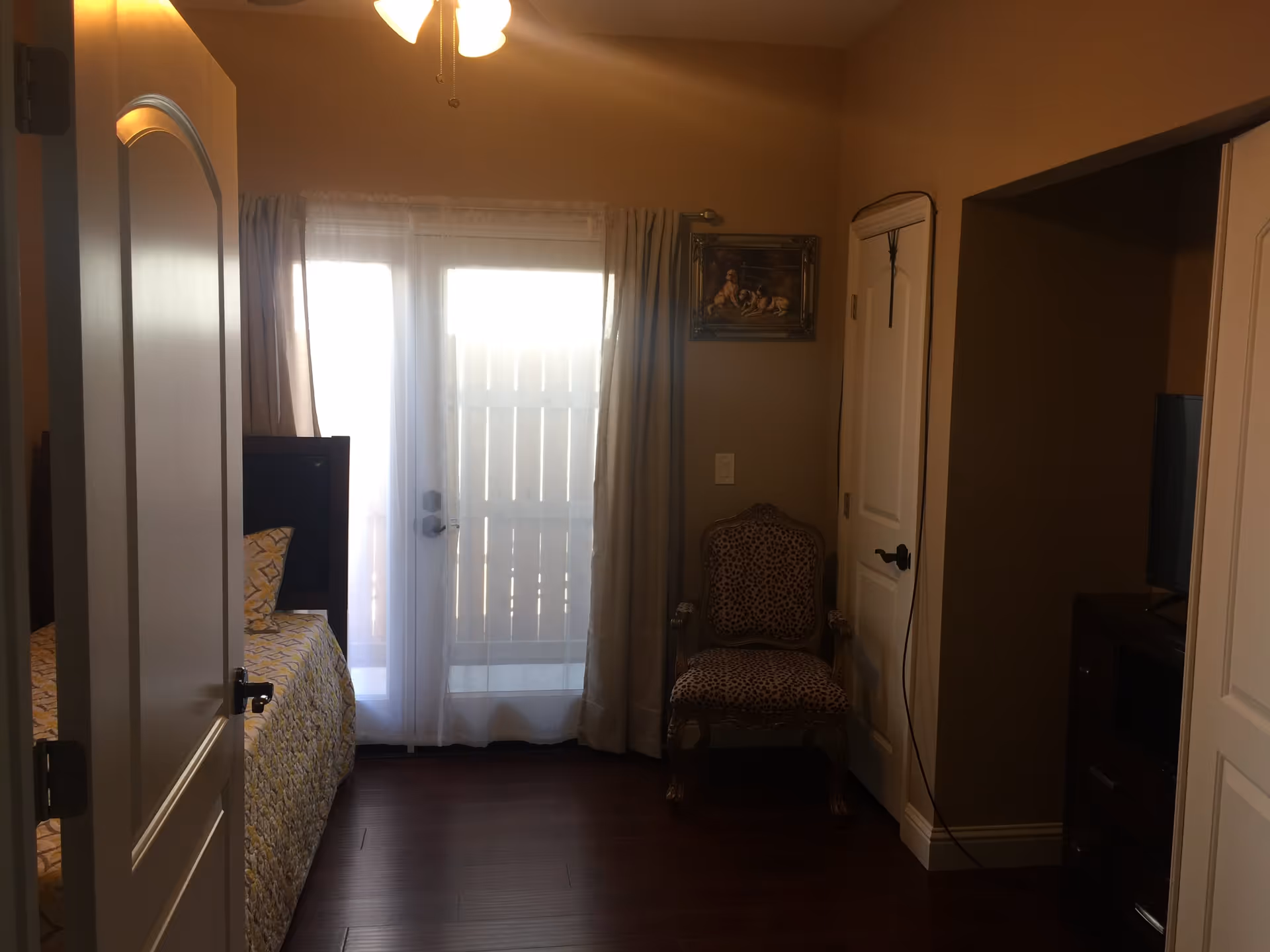 A bedroom with a partially open door showing a bed with a yellow patterned bedspread and pillow. There is a glass door with sheer curtains letting in natural light. A leopard print upholstered chair is placed against the wall next to a closed white door. A small framed painting hangs above the chair. A television is visible on a dark wooden dresser to the right.