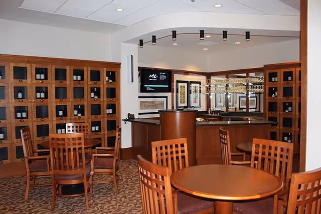 Interior view of a lounge area with wooden tables and chairs, a bar counter with glass shelves and wine bottles displayed in wooden cubbies along the wall. The ceiling has recessed lighting and there is a TV mounted on the wall.