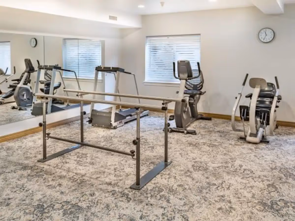 Exercise/therapy room with parallel bars, treadmill and stationary bikes in front of a mirrored wall.