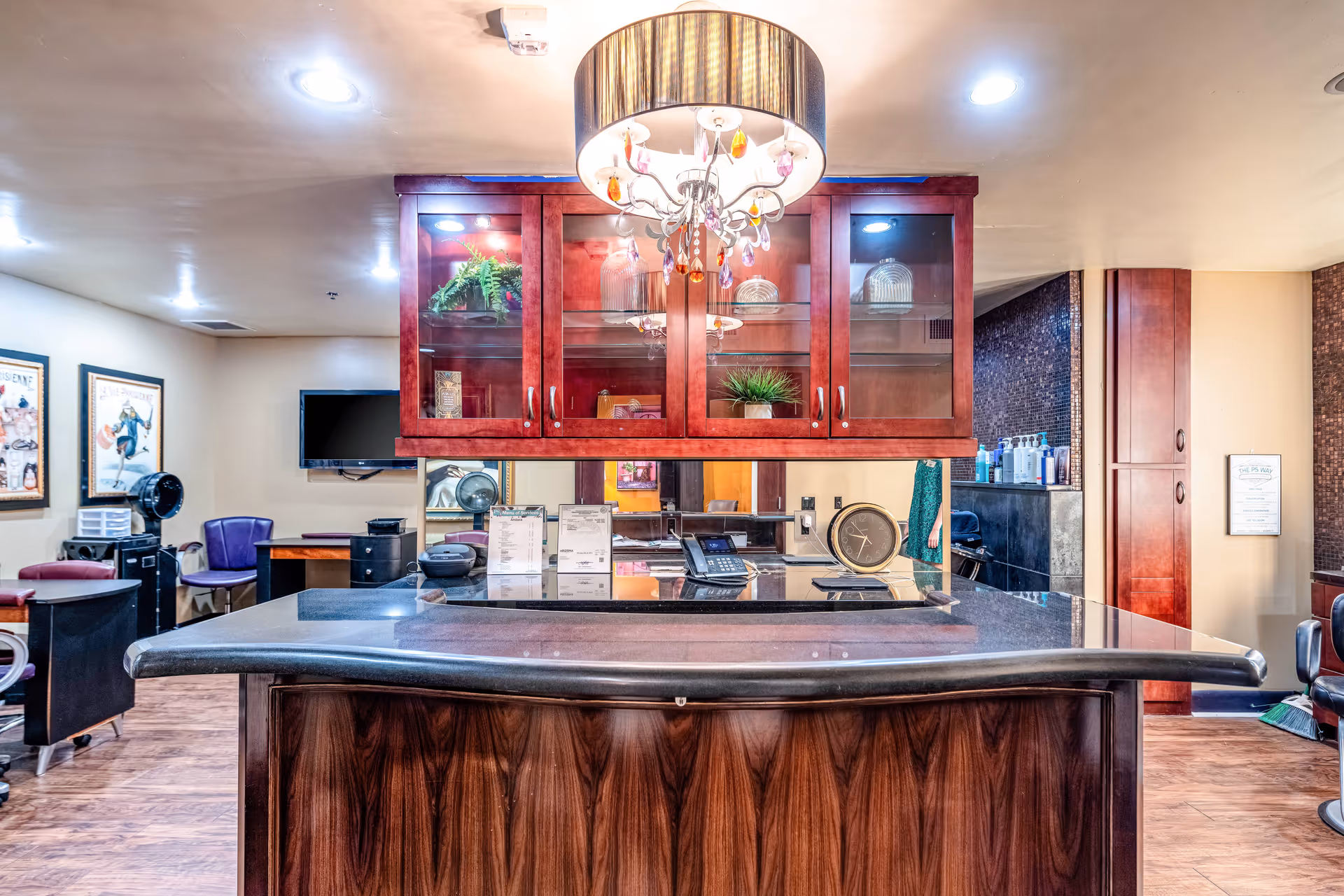 Interior view of a reception or front desk area with a dark wood counter and a hanging cabinet above it. The cabinet has glass doors displaying decorative items and plants. A chandelier with hanging crystals is suspended from the ceiling above the counter. In the background, there are chairs, framed artwork on the walls, a wall-mounted TV, and various office equipment.