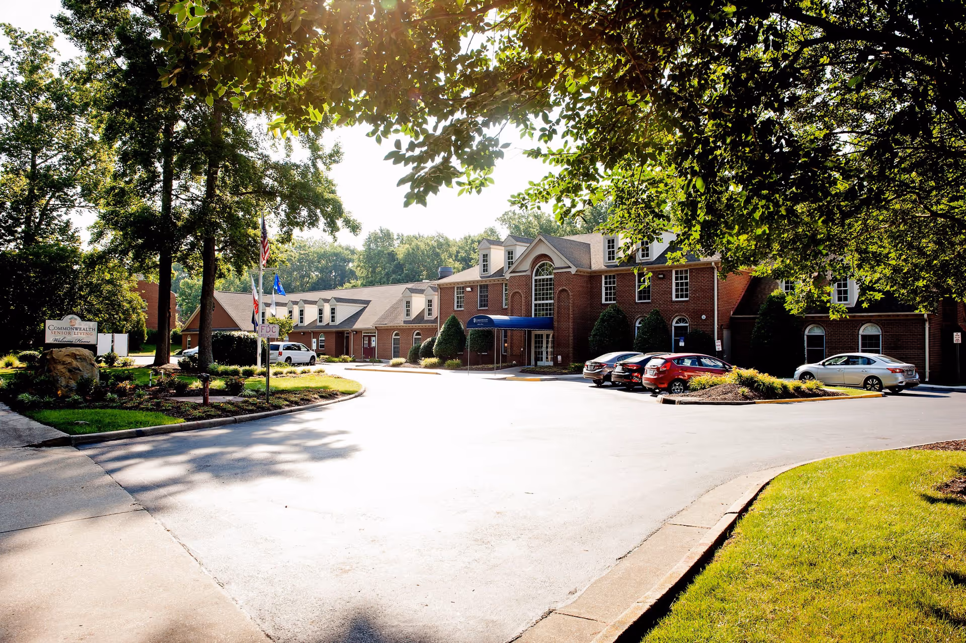 Exterior view of Commonwealth Senior Living at Georgian Manor, showing a large brick building with multiple windows and a blue awning over the entrance. There are several cars parked in front, a well-maintained driveway, and green trees and landscaping surrounding the area.