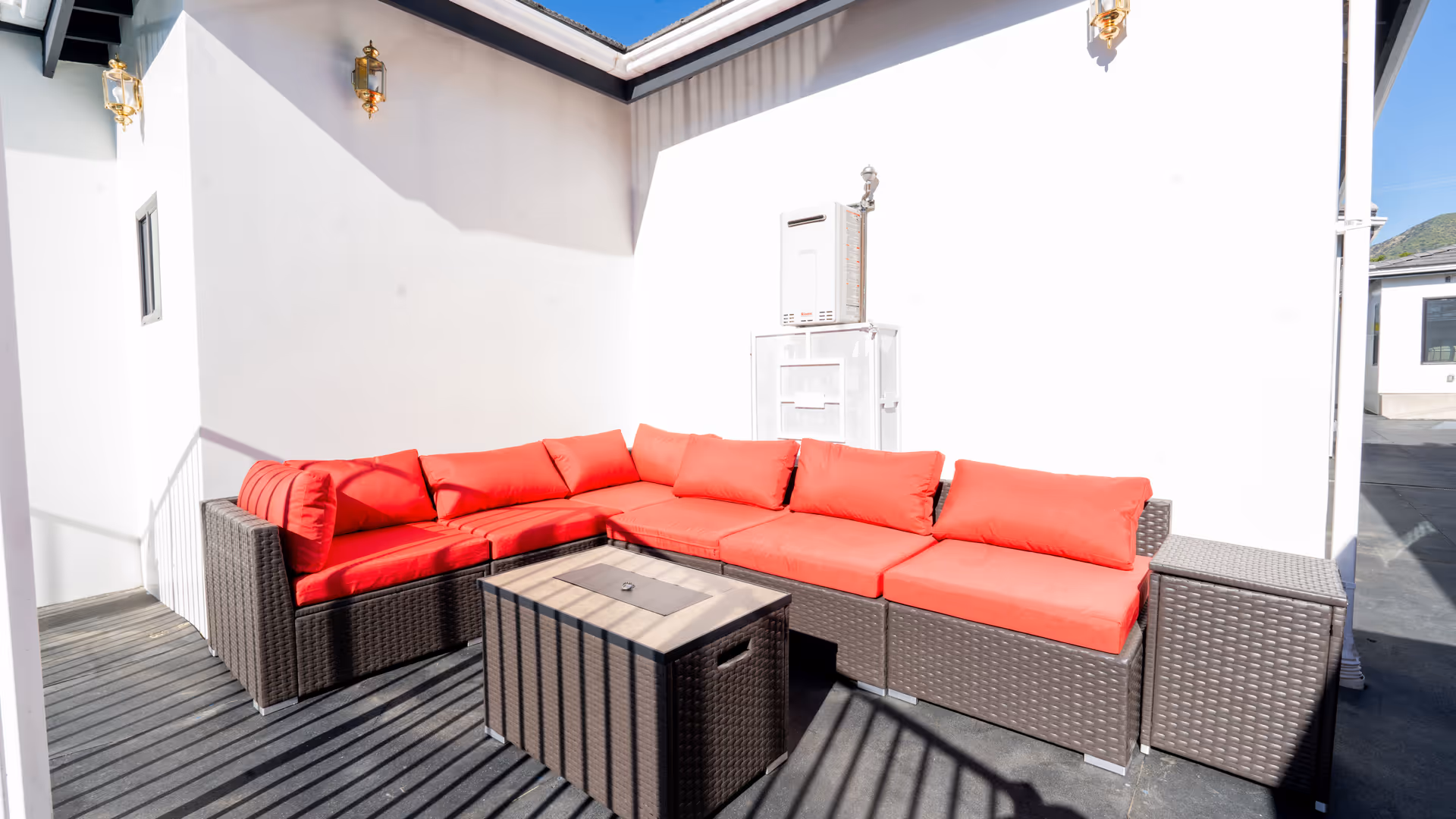 Outdoor patio area with a brown wicker sectional sofa featuring bright red cushions and a matching wicker coffee table. The space is enclosed by white walls with two gold wall-mounted lanterns and a clear blue sky above.