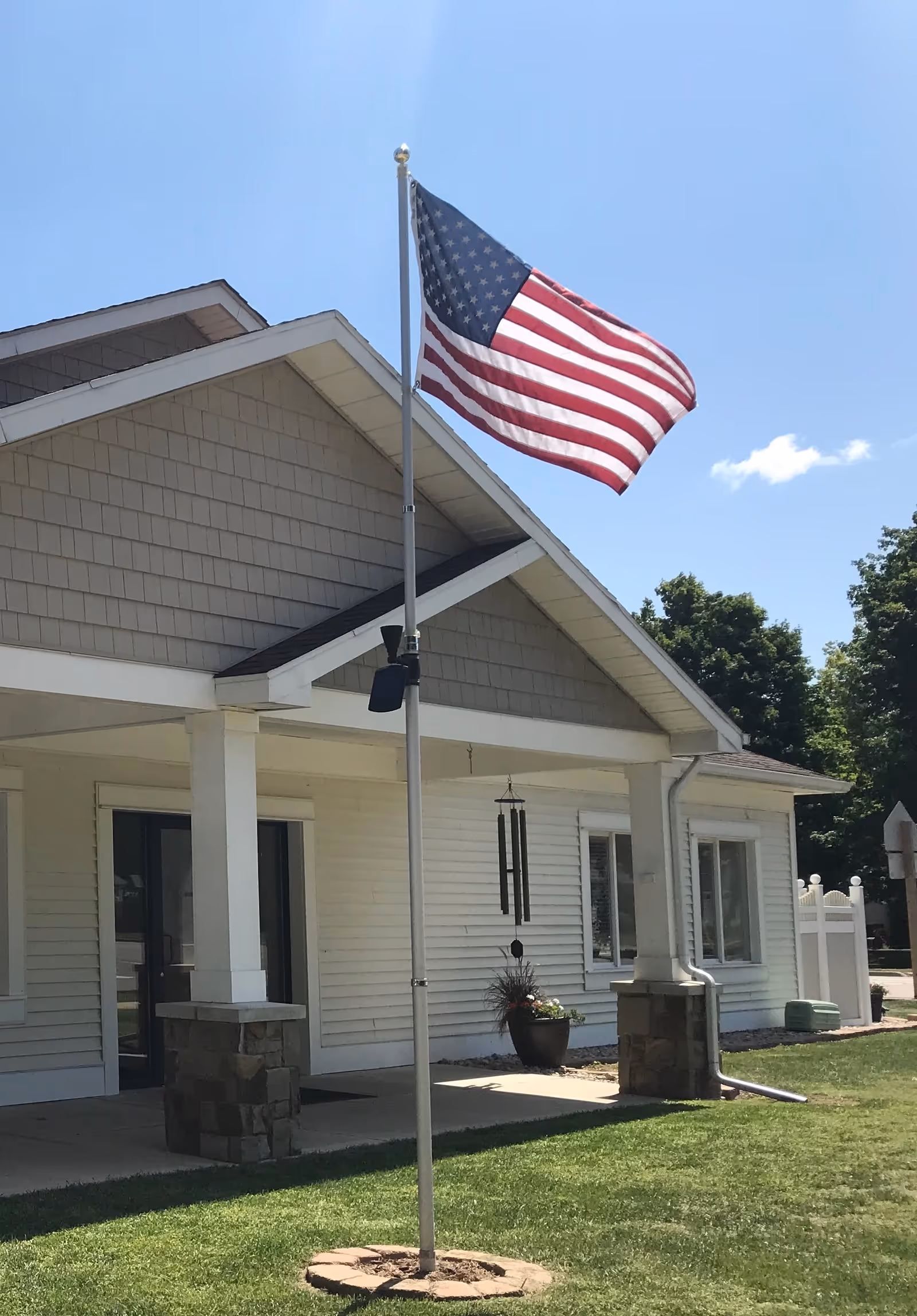 Exterior view of a single-story building with white siding and a covered porch supported by white columns with stone bases. An American flag is flying on a flagpole in front of the building, and there is a wind chime hanging near the entrance. The sky is clear and blue with some trees in the background.
