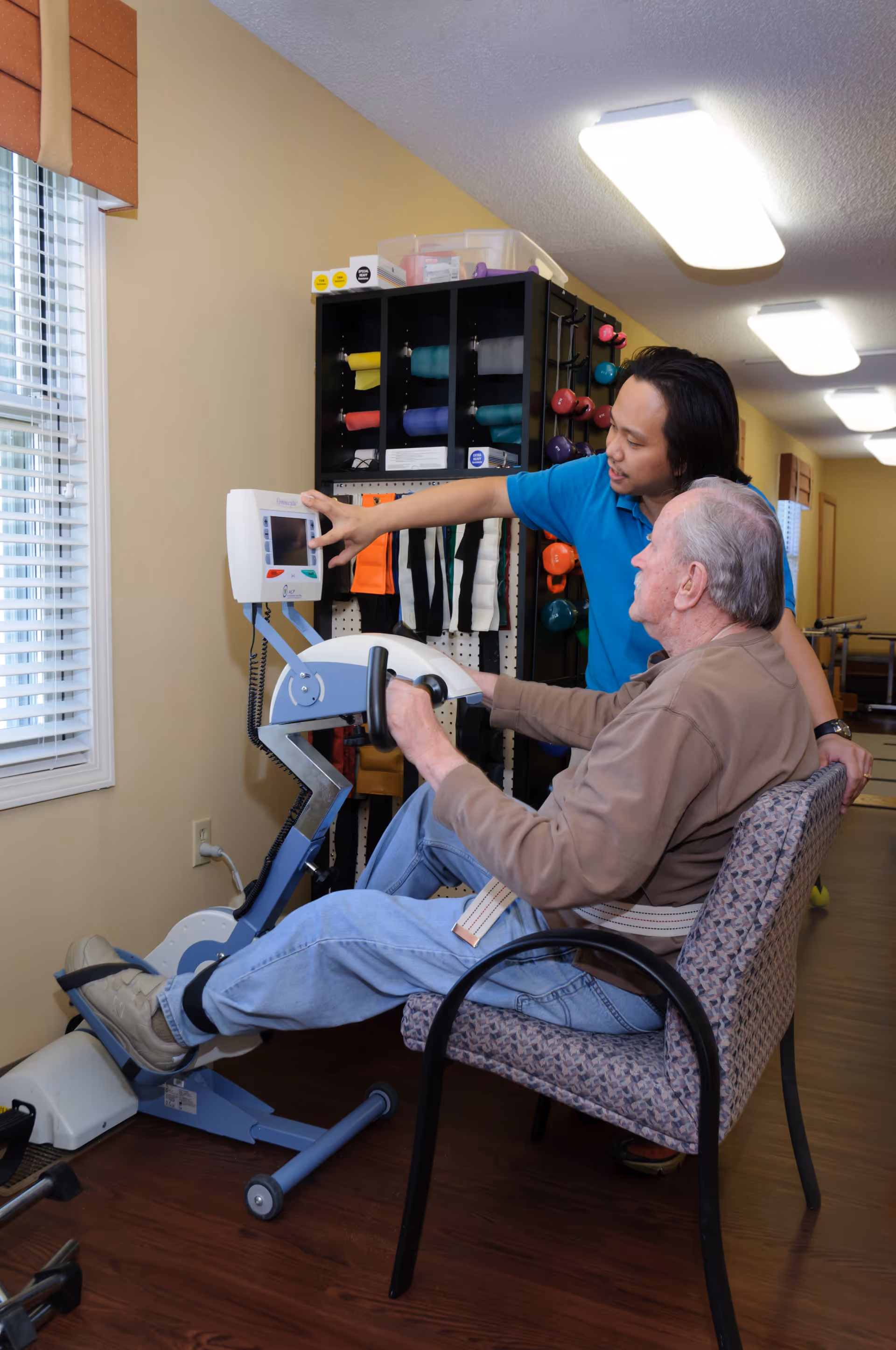 An elderly man seated on a chair using a pedal exercise machine while a caregiver in a blue shirt assists him by pointing at the machine's display. The room has a window with blinds, shelves with colorful exercise bands and weights, and overhead fluorescent lighting.