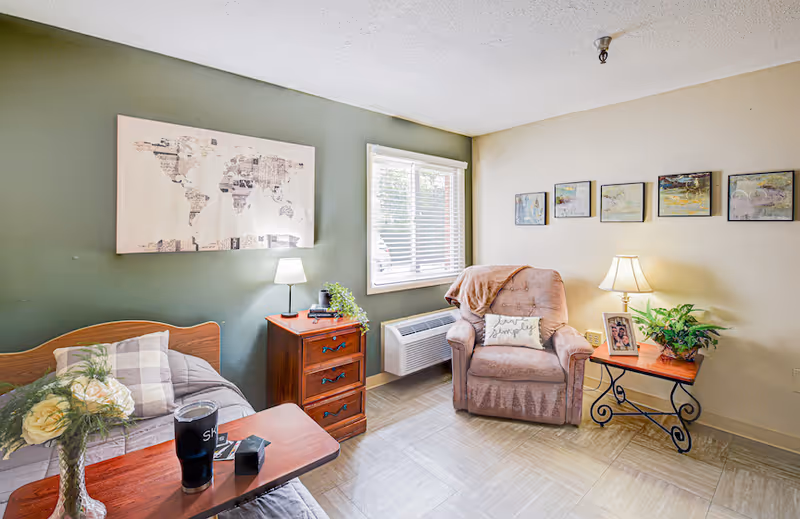 Small furnished senior living bedroom with a bed, bedside tables, an armchair, wall art and a window.