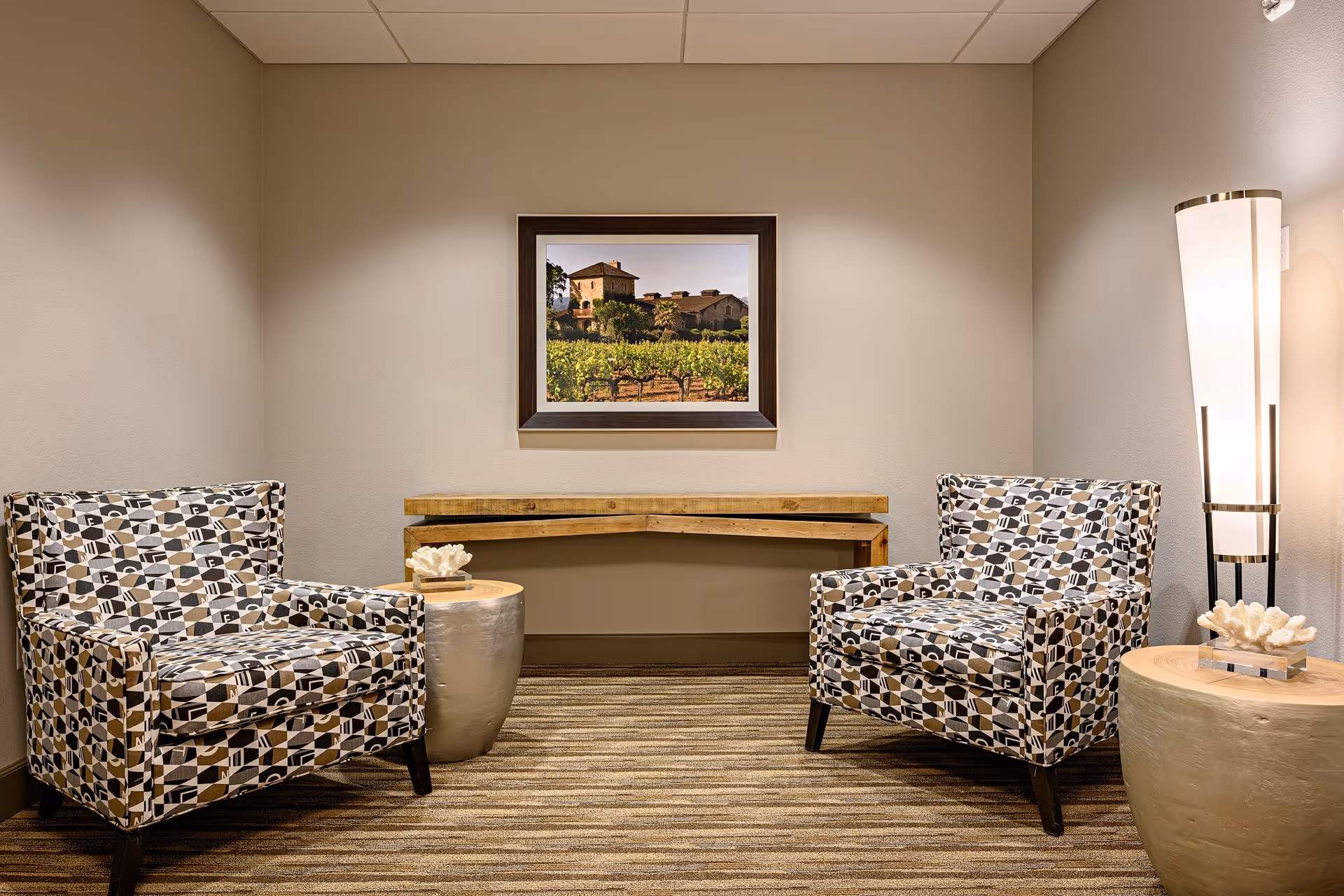 A cozy seating area with two patterned armchairs facing each other, a wooden corner table mounted on the wall, two round side tables with decorative coral pieces, and a tall modern floor lamp. A framed picture of a vineyard and building hangs on the beige wall behind the table.