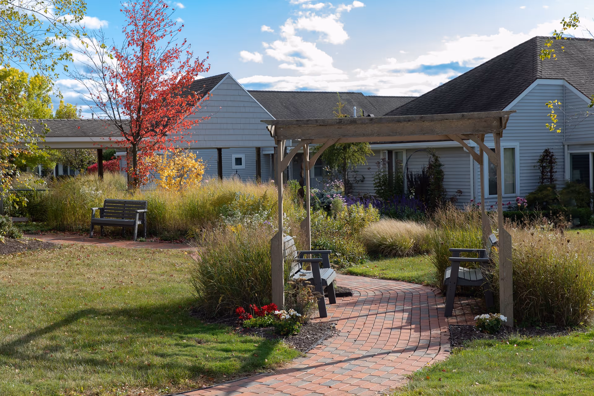 A peaceful outdoor garden area at Kendal at Ithaca featuring a curved brick pathway leading under a wooden pergola with benches on either side. The garden is surrounded by tall grasses, colorful flowers, and trees with autumn foliage. Residential buildings with gray siding and dark roofs are visible in the background under a partly cloudy sky.