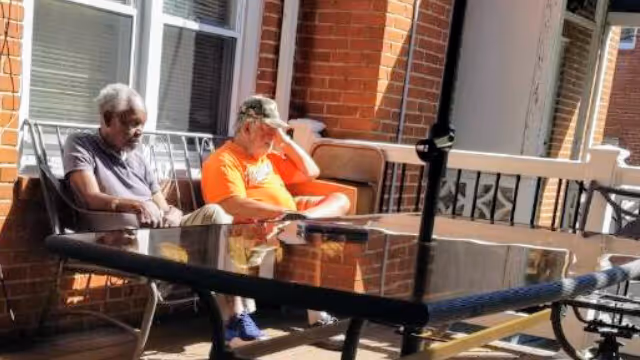 Two elderly men sitting on a porch or patio area with brick walls and windows behind them. One man is wearing a gray shirt and beige pants, and the other is wearing an orange shirt and a camouflage cap. There is a glass-top table with a black frame in front of them, and a black umbrella pole is visible in the center of the table.