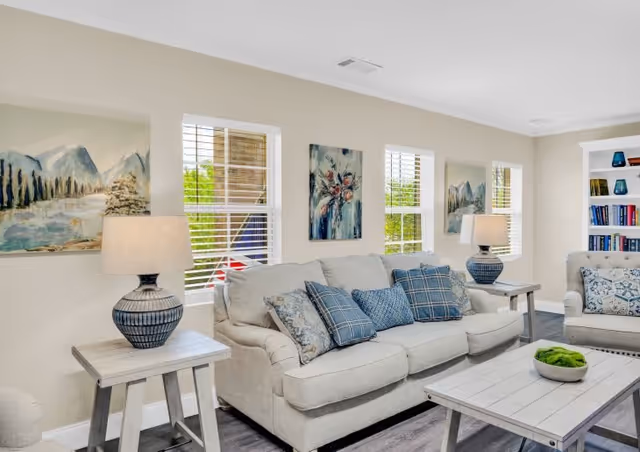 A bright living room with a beige sofa and armchair adorned with blue and patterned cushions. Two matching table lamps sit on side tables next to the sofa. The walls are decorated with three paintings, and there are three windows with white blinds letting in natural light. A white wooden coffee table with a green decorative bowl is in front of the sofa, and a built-in bookshelf with books and decorative items is visible in the corner.