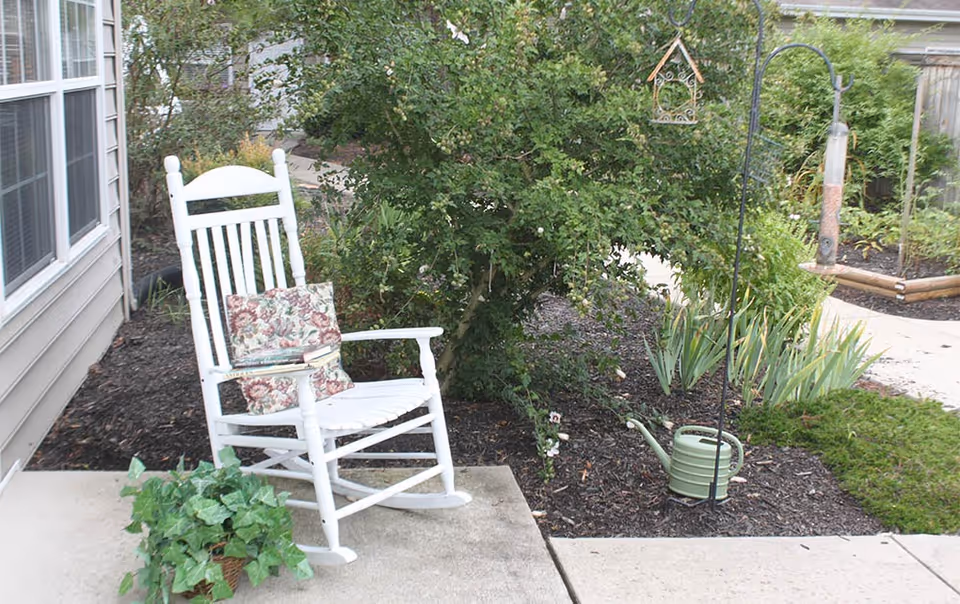 White wooden rocking chair with floral cushions placed on a concrete patio next to a house. Surrounding the chair are garden plants, a green watering can, and a bird feeder hanging from a metal hook. The area is landscaped with mulch and greenery.
