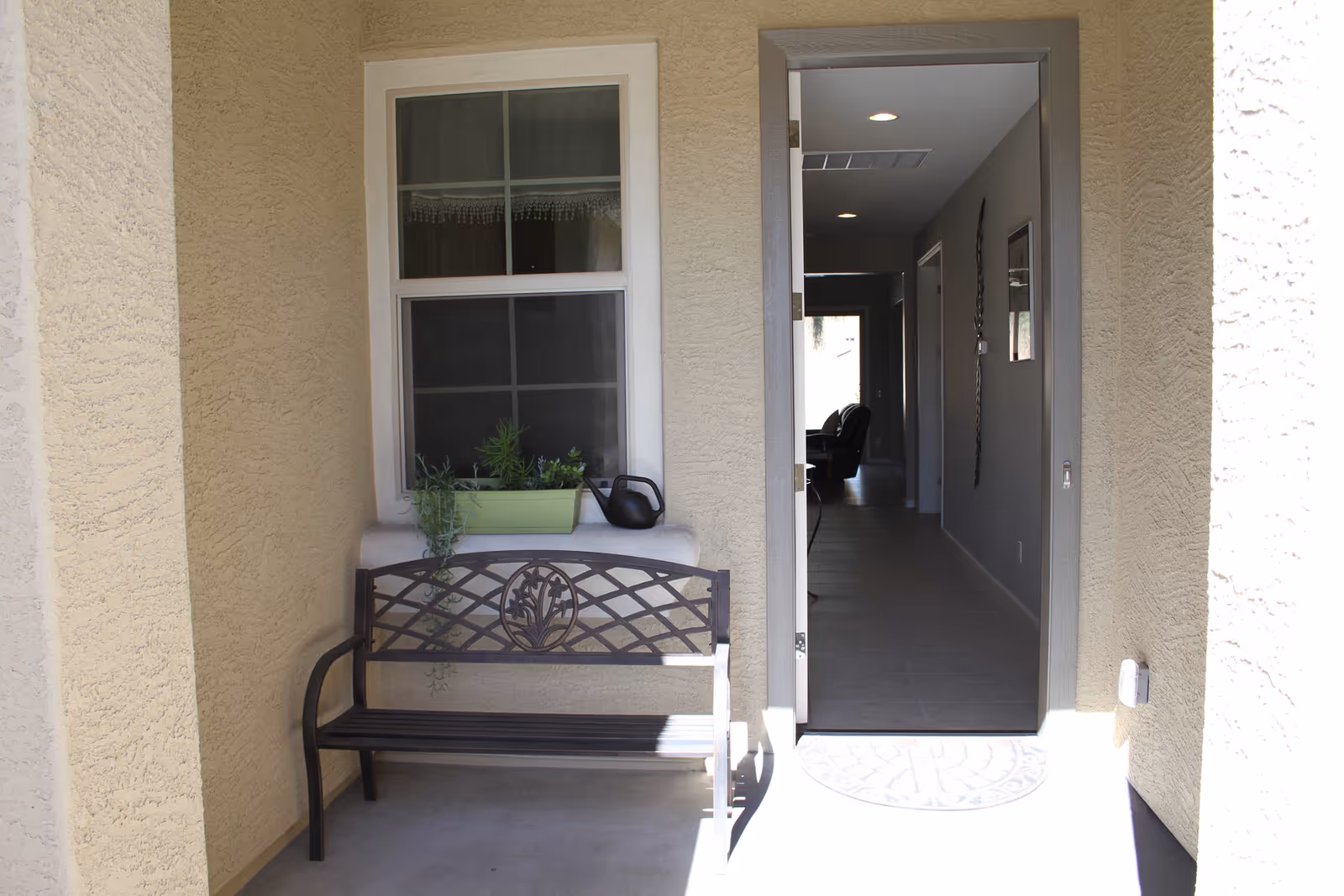 A small outdoor porch area with a decorative metal bench placed against a textured beige wall. Above the bench is a window with a green planter box containing plants and a black watering can. To the right, an open door leads into a hallway with tiled flooring and recessed ceiling lights, with a glimpse of a chair further inside.