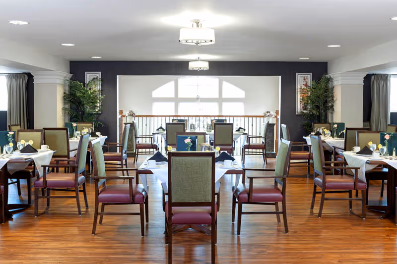 A bright and spacious dining room with multiple tables set for meals. Each table is covered with a white tablecloth and has chairs with green backs and red seats. The room features large windows letting in natural light, wooden flooring, decorative plants, and modern ceiling lights.