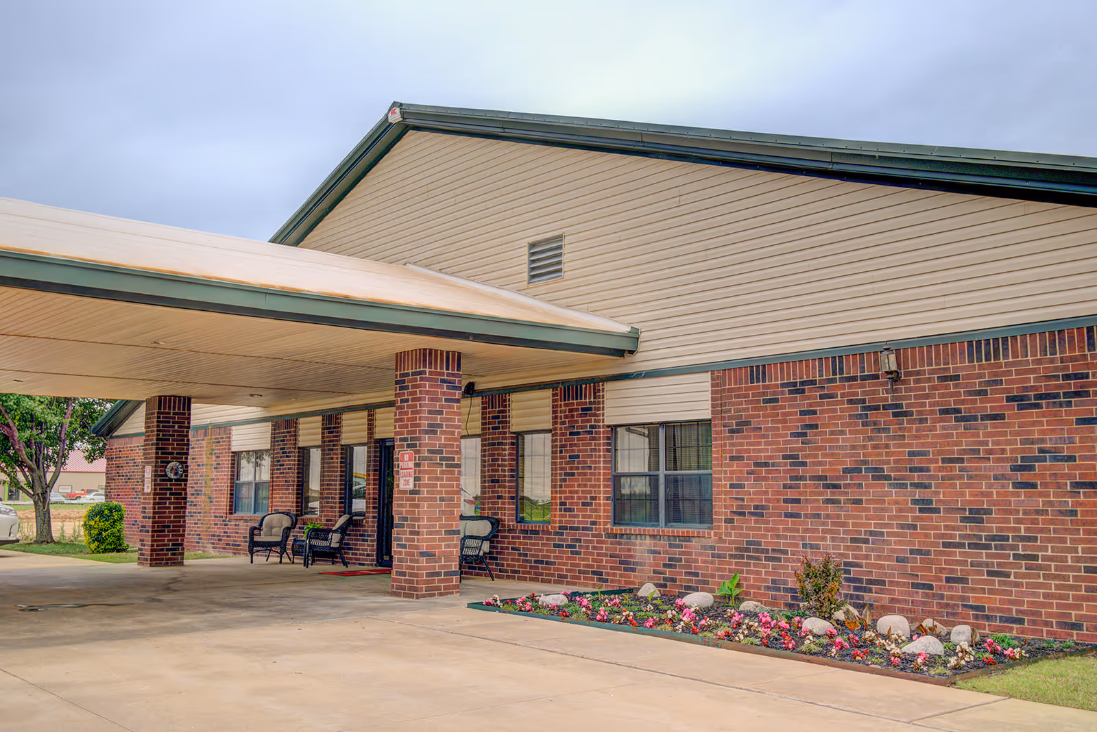 Exterior view of a brick and siding building with a covered entrance supported by brick pillars. There are chairs placed near the entrance and a flower bed with colorful flowers along the side of the building.