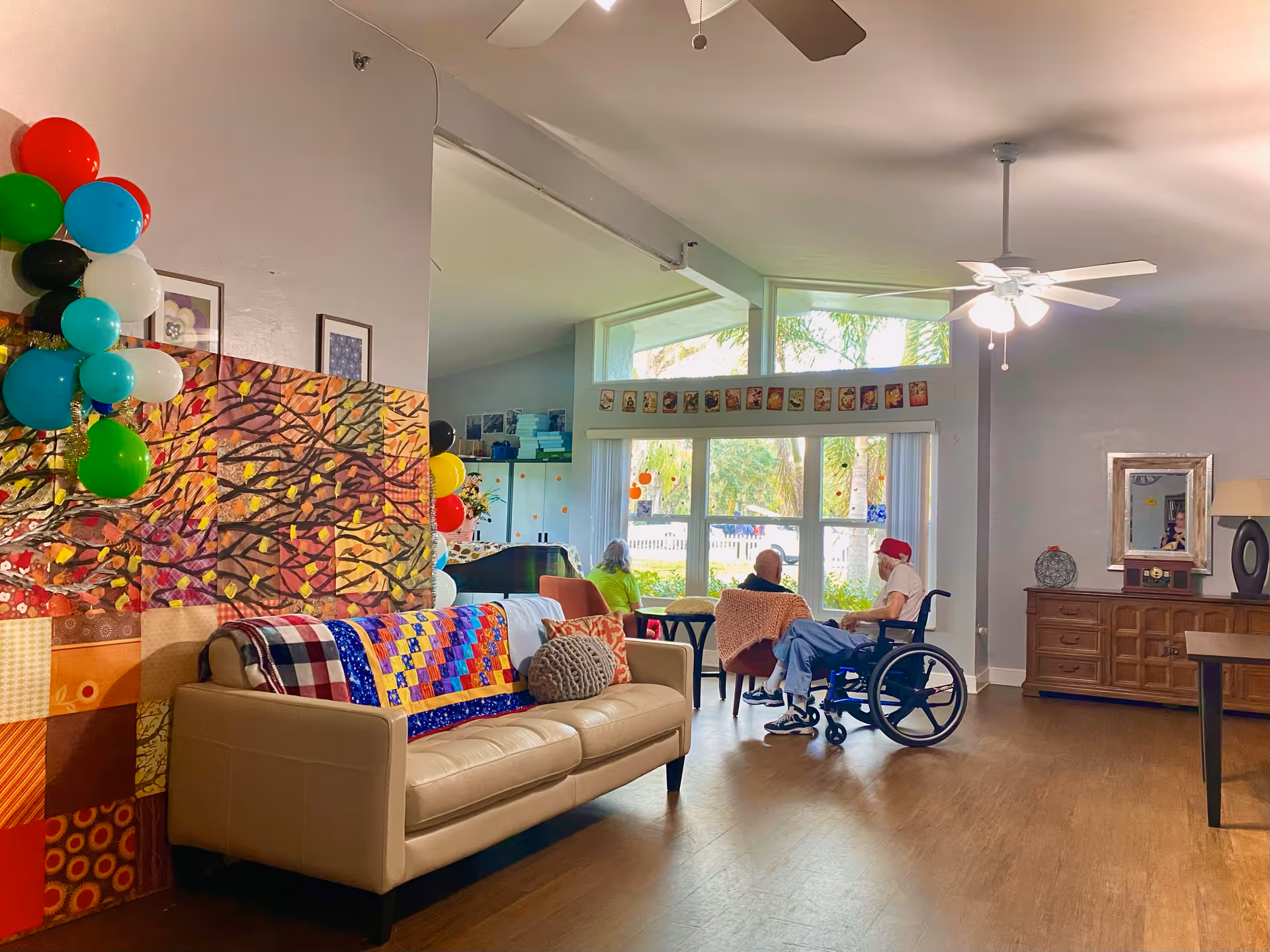 A bright and cozy common area in a senior living facility with a beige couch adorned with colorful quilts and pillows. Three elderly people are seated near large windows that let in natural light, with one person in a wheelchair. The room features wooden flooring, a ceiling fan, colorful balloons, and decorative artwork on the walls.