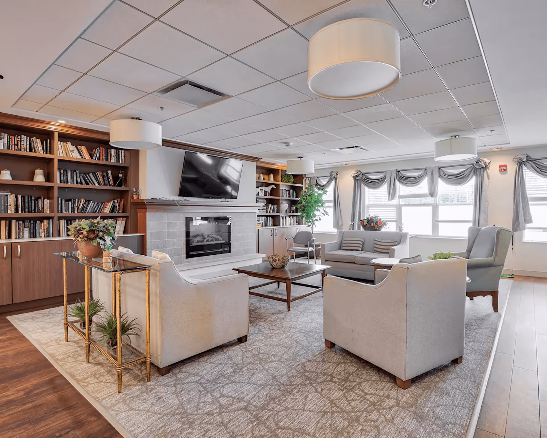 A bright and spacious living room area with comfortable seating including two armchairs and a sofa arranged around a wooden coffee table. The room features a fireplace with a mounted flat-screen TV above it, built-in wooden bookshelves filled with books and decorative items, large windows with gray curtains allowing natural light to fill the space, and modern ceiling lights. A small table with plants is positioned behind one of the armchairs, and the floor is covered with a patterned area rug over wood flooring.