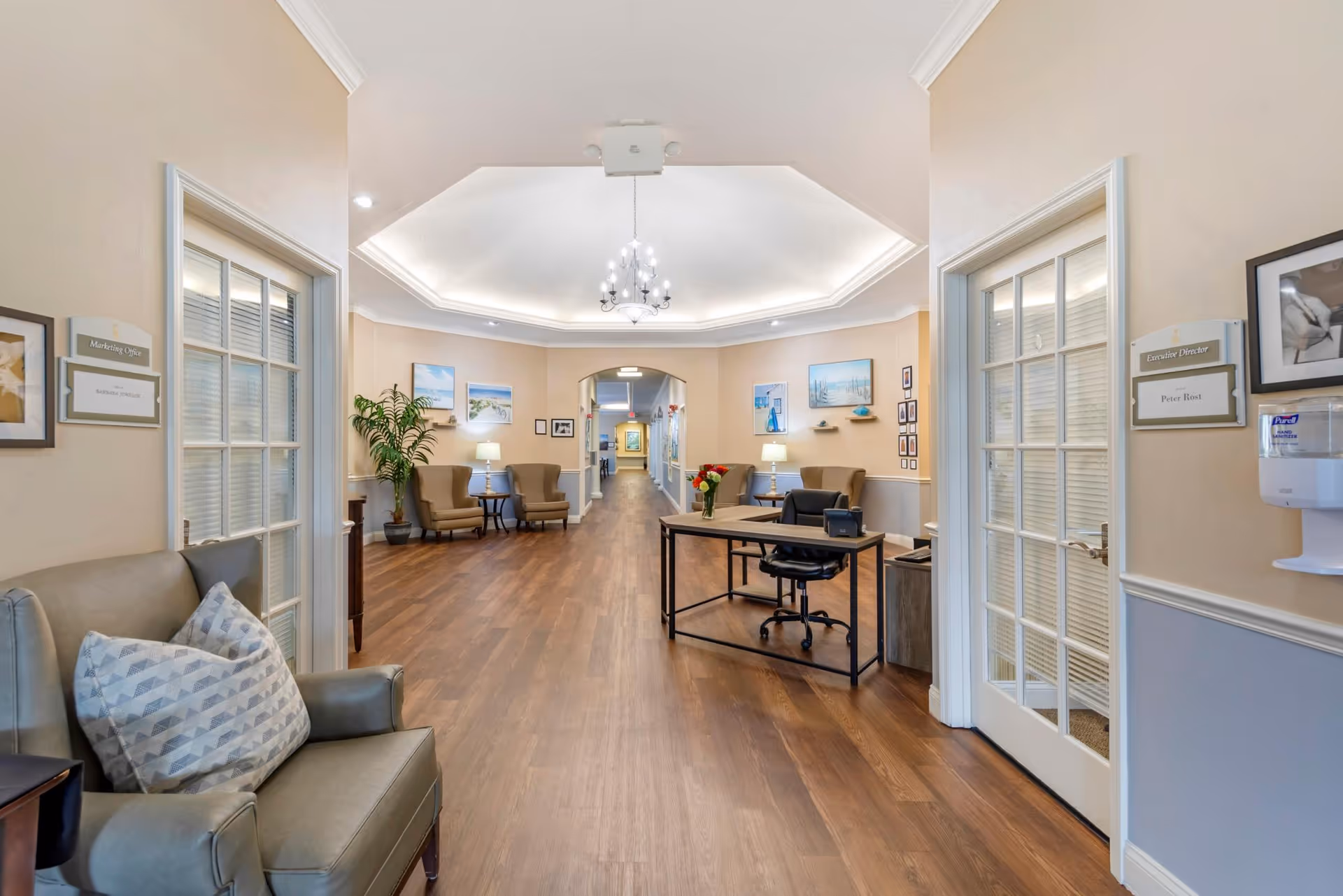 Bright, welcoming senior living facility lobby with seating, a reception desk, and a long wood-floored hallway.