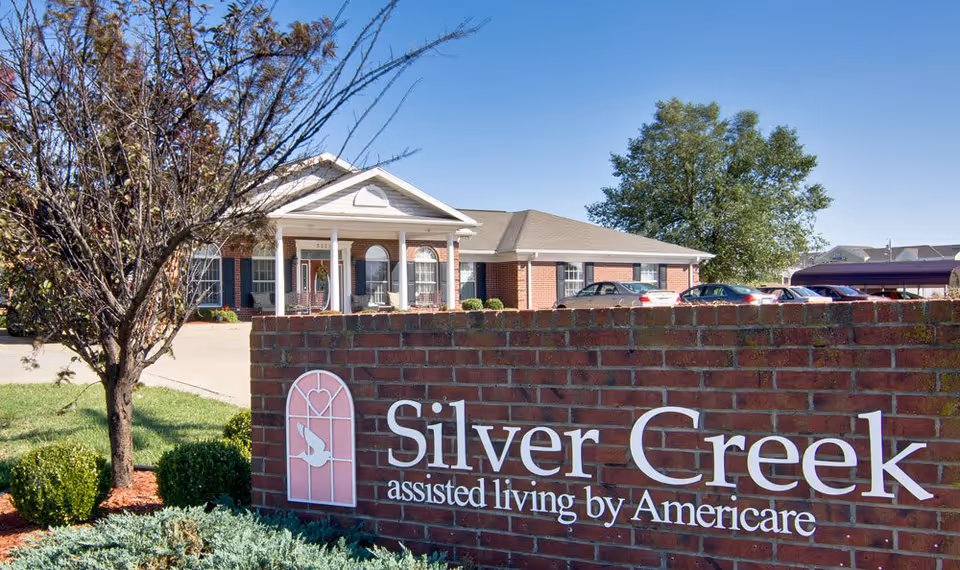 Exterior view of Silver Creek assisted living facility showing a brick sign with the facility name and a building with a porch and columns in the background under a clear blue sky.