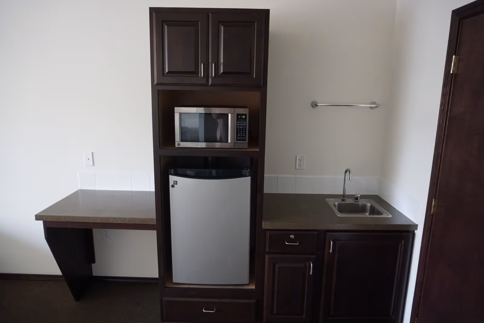 A small kitchenette area with dark wood cabinets, a microwave, a mini refrigerator, a countertop with a sink, and an empty towel rack mounted on the wall. There is also a small desk area attached to the left side of the kitchenette.