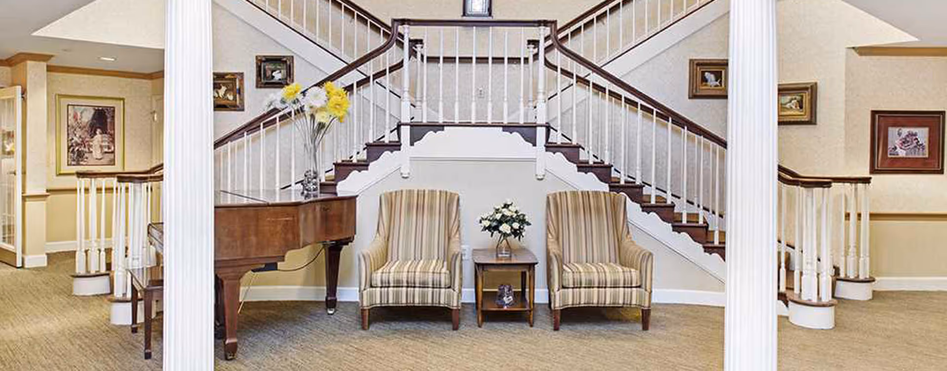 Lobby with a grand piano, two striped armchairs flanking a small table and a double staircase in the background.