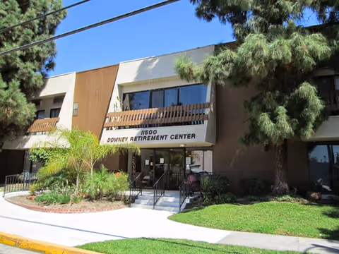 Exterior view of Downey Retirement Center building with a brown and white facade, large windows, and an entrance with steps and railings. There are trees and landscaped greenery around the building under a clear blue sky.