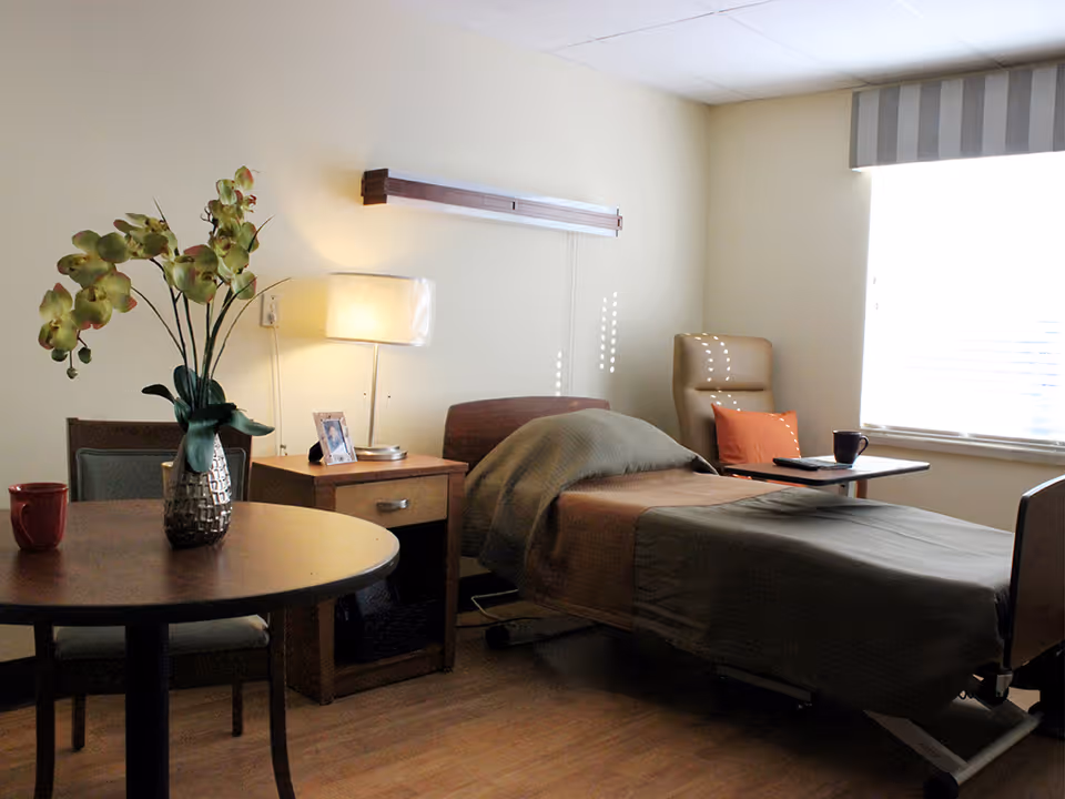 A cozy bedroom in a senior living facility with a single bed covered in a green blanket, a wooden nightstand with a lamp and a framed photo, a round table with a vase of flowers and a red mug, and a comfortable chair with an orange pillow near a window with blinds.