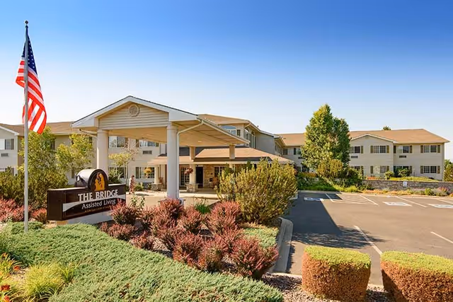 Exterior view of The Bridge Assisted Living facility with a covered entrance, landscaped bushes, an American flag on a flagpole, and a clear blue sky.