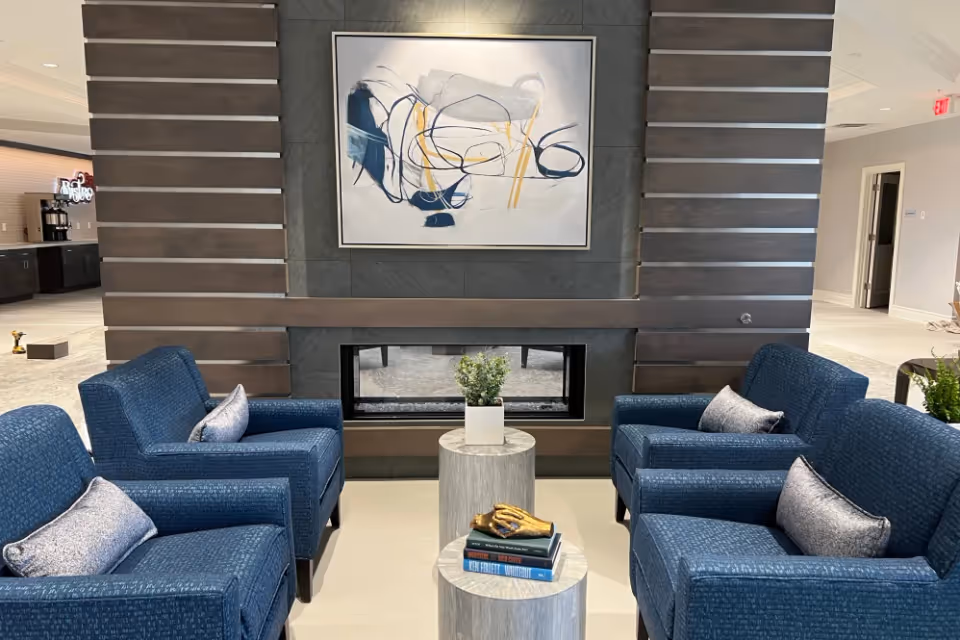 Lobby seating area with four blue armchairs arranged around two round tables in front of a modern double-sided fireplace and abstract painting.