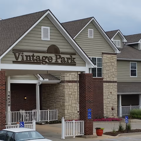 Entrance of a building with a sign that reads Vintage Park. The building features a combination of brick and stone exterior with green siding and white trim. There are handicap parking signs and a parked car visible in front.