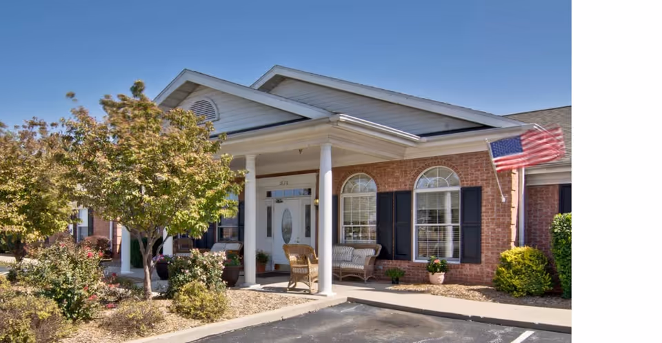 Front entrance of an assisted living building with a covered porch, columns, seating, landscaped beds, and an American flag.