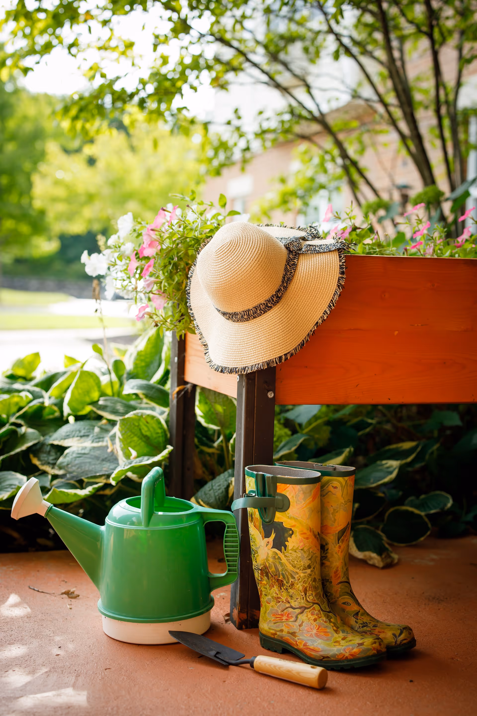 A pair of colorful floral rain boots, a green watering can, a small garden trowel, and a straw hat hanging on the edge of a wooden planter box filled with blooming flowers, set on a patio with green leafy plants and trees in the background.